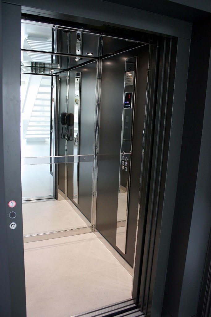 Inside the dark custom lift cabin with stainless steel trim, a control panel and glass doors opening to a white landing