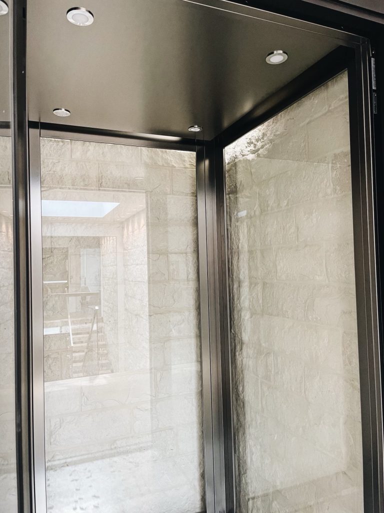 Looking through the glass of the E1 lift beside the black-framed wine display, with the stone shaft wall behind