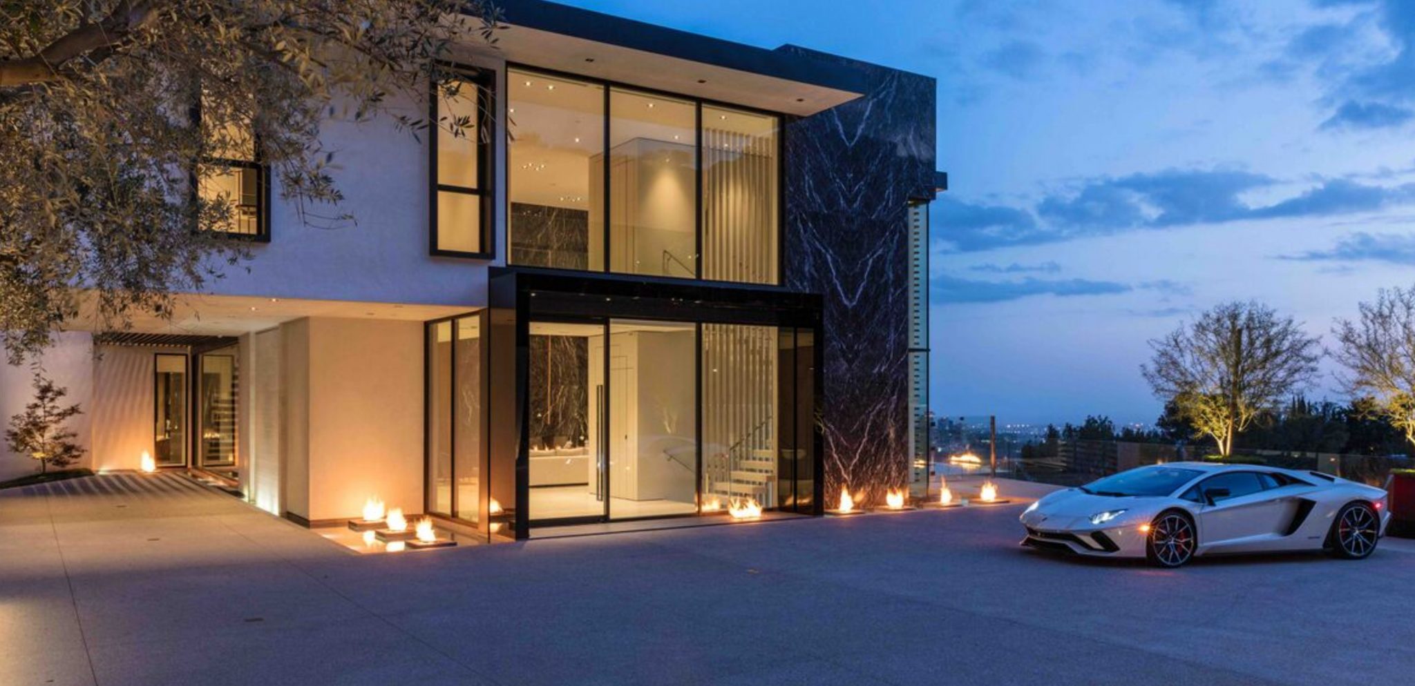 Modern two-storey Los Angeles estate at dusk with a glass and black marble facade, lit fire features and a white sports car on the driveway