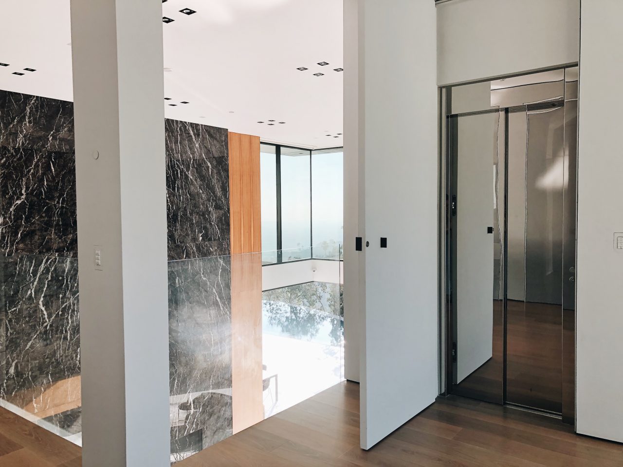 E2 home lift with a polished stainless steel door beside a black marble wall and a white pivot door, with the infinity pool below