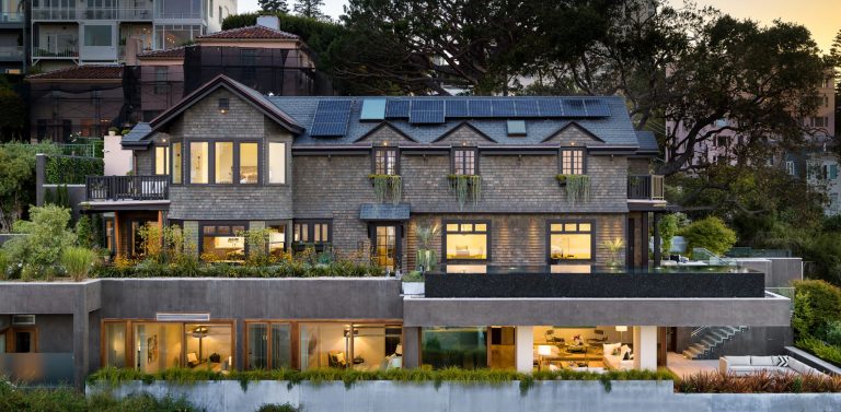 House with a shingle clad roof and glowing windows at dusk set among terraced gardens