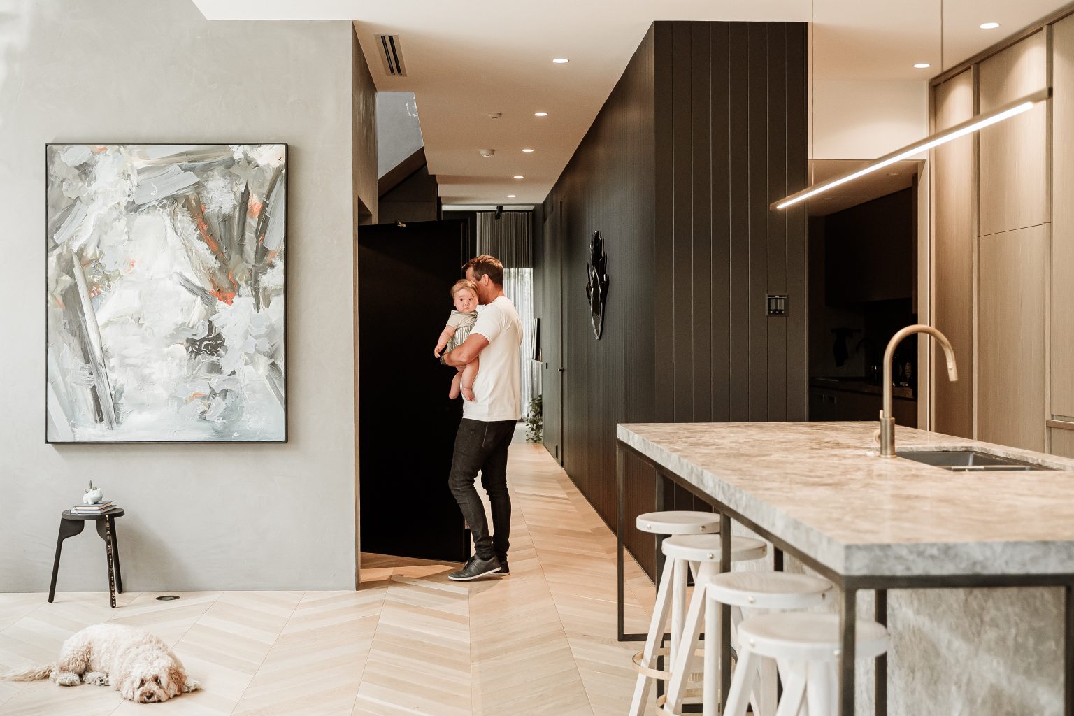 Father carrying his baby to the lift door past the kitchen island, with the family dog resting on the herringbone floor