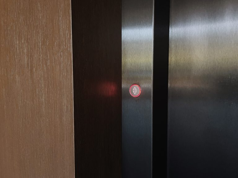 Close view of a dark steel lift door with a red ring call button beside a timber wall panel