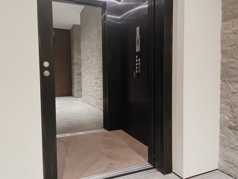 Open dark framed lift door showing the button panel and chevron-pattern floor beside a stone wall
