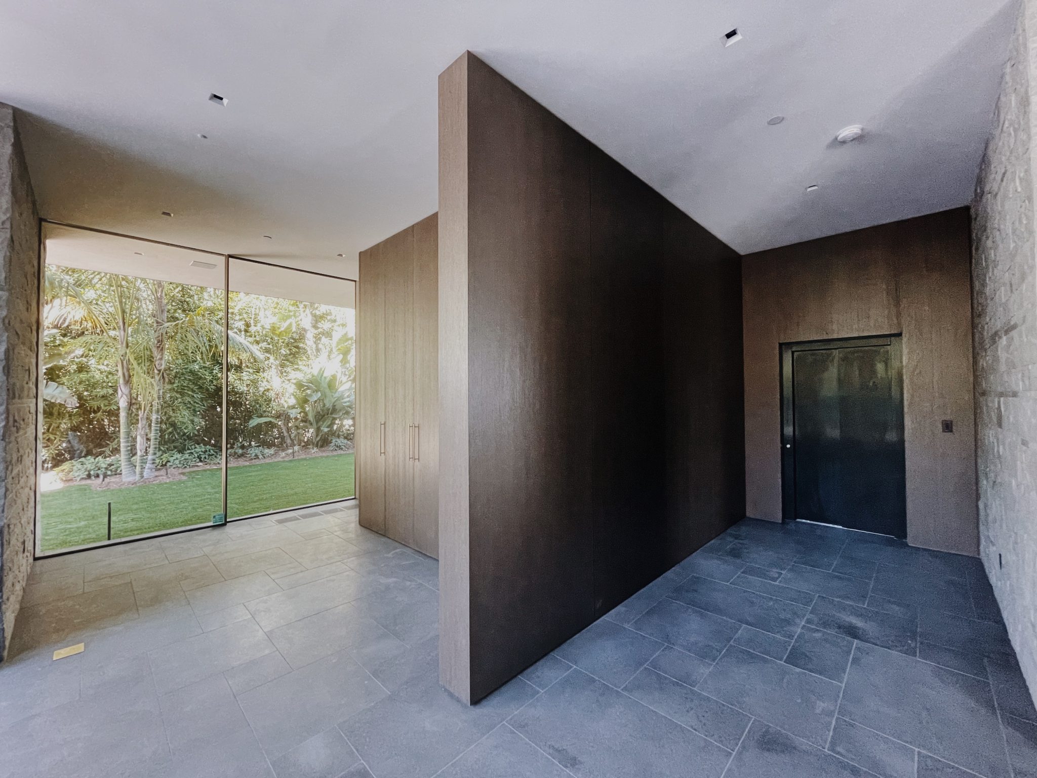 Slate-floored lift lobby with a timber wall panel, a dark lift door and a glass wall to the garden in a Los Angeles hillside home
