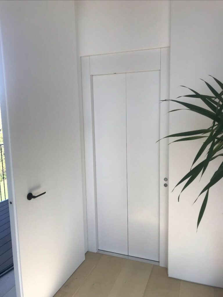 Closed white lift doors set flush in a white wall beside a potted plant