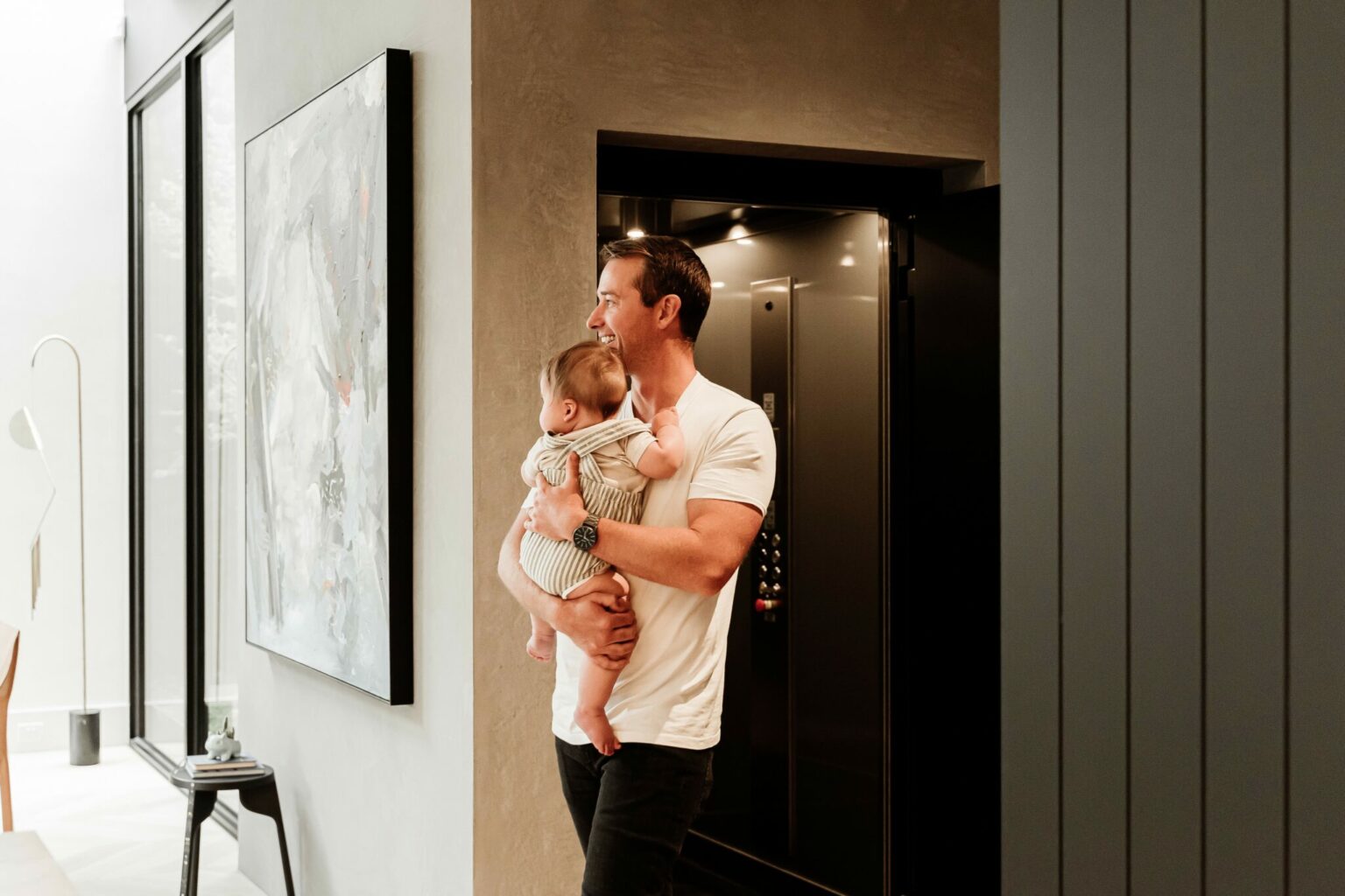 Father holding his baby beside the open E1 swing door lift in a Melbourne home, with the brushed stainless steel cabin behind him