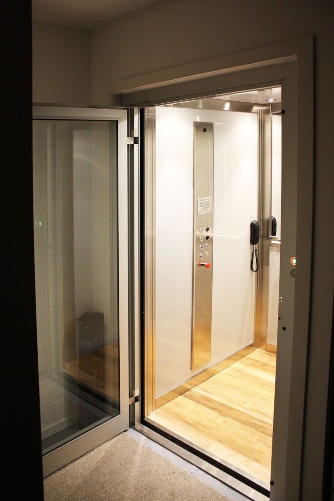 The lift landing door open on the upper floor in the evening, the lit cabin inside