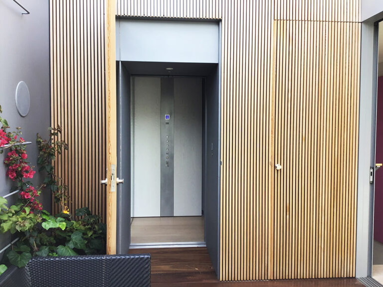Entry with timber battens, a flush door and the lift door framed by plants in Vaucluse
