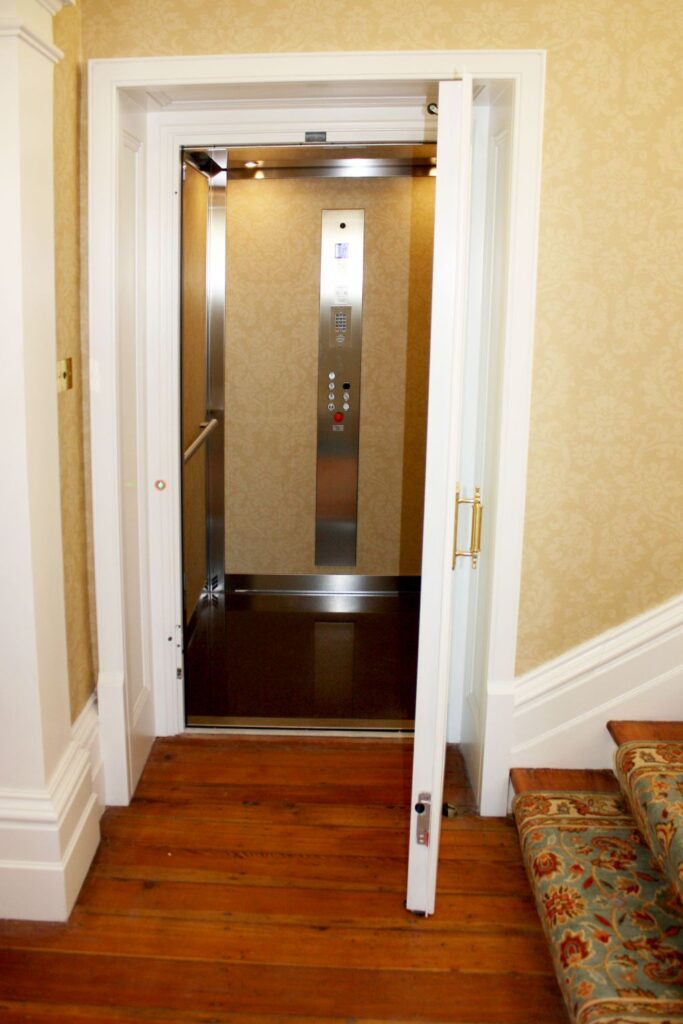 Open white panelled door showing the E1 lift cabin with a timber floor beside a patterned stair runner