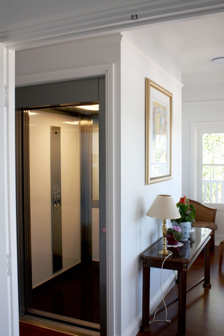 Open stainless steel lift door on an upstairs landing of the Queenslander, beside a timber side table, lamp and framed artwork