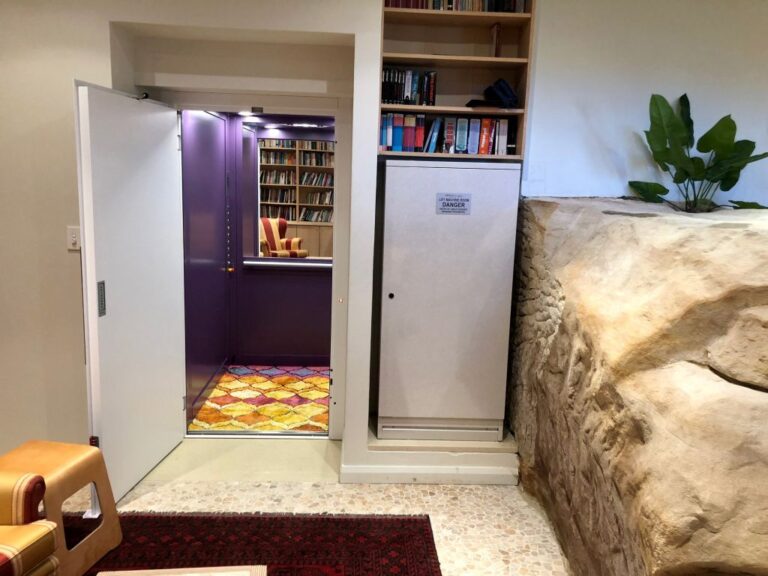 Lift door open to a purple cabin beside a bookshelf and a sandstone look rock feature
