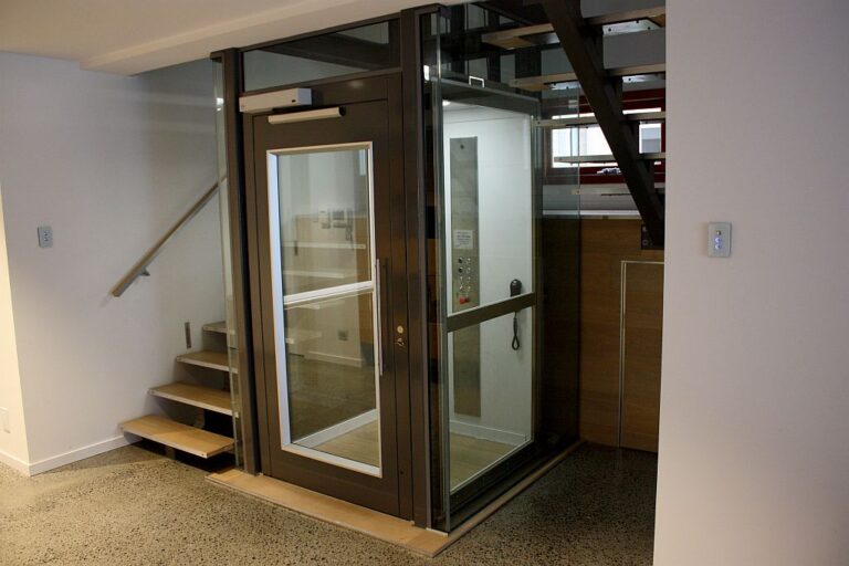 Dark framed home lift with glass panel beside a timber staircase and polished concrete floor