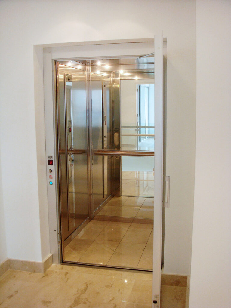 ElfoLift in its glass shaft with downlights and a stone tile floor, seen from the front