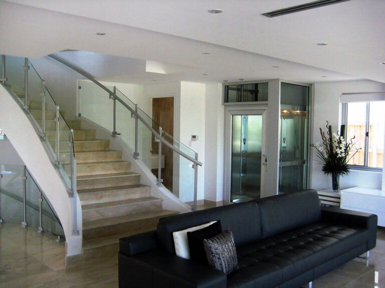 Curved staircase with glass balustrade beside a glass lift and a black leather sofa