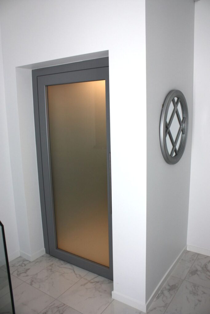 Closed E1 lift landing door with a grey frame and frosted glass in a white wall beside a round decorative mirror