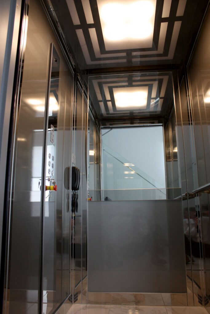 Inside the lift car looking up at the patterned ceiling light, with polished stainless steel walls and car operating panel