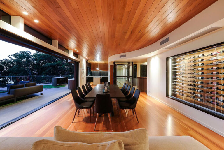 Timber ceilinged dining room with a long table, a wine wall and the glass home lift in the background