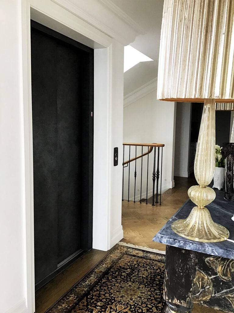 Closed dark lift door beside a lamp and a curved staircase balustrade in a Sydney home
