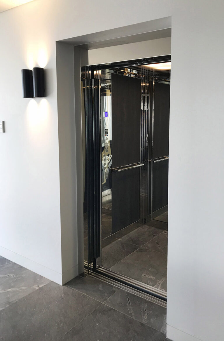Open lift doorway in a white wall beside a black wall light, showing the dark panelled cabin with mirrored walls and polished stainless steel trim