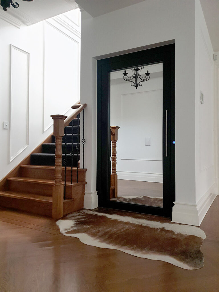 Closed E1 lift door with a full-height mirror panel beside the timber staircase and chandelier in Eaglemont