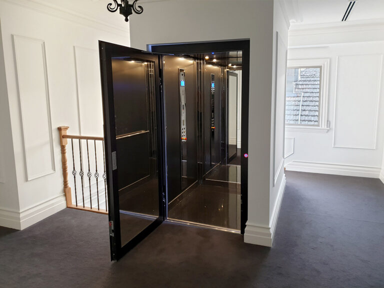 Open black-framed swing door E1 lift on the upstairs landing with grey carpet and a white panelled wall