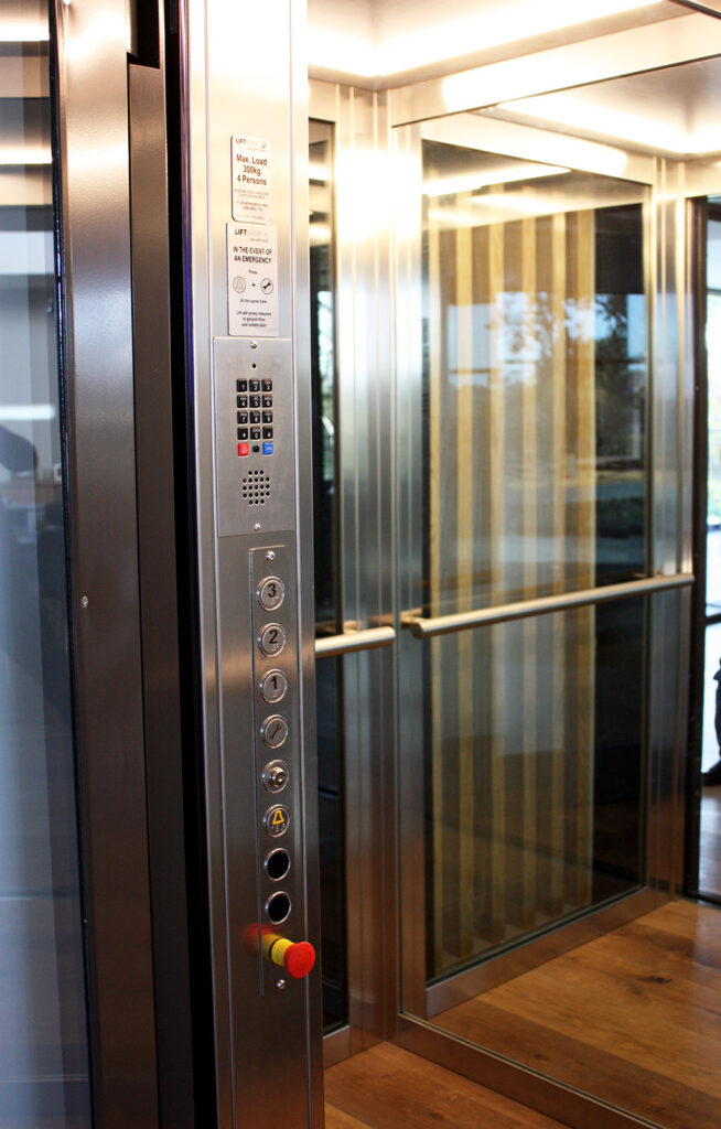 Inside the E1 lift cabin with brushed stainless control panel, handrail and glass and gold-toned panels
