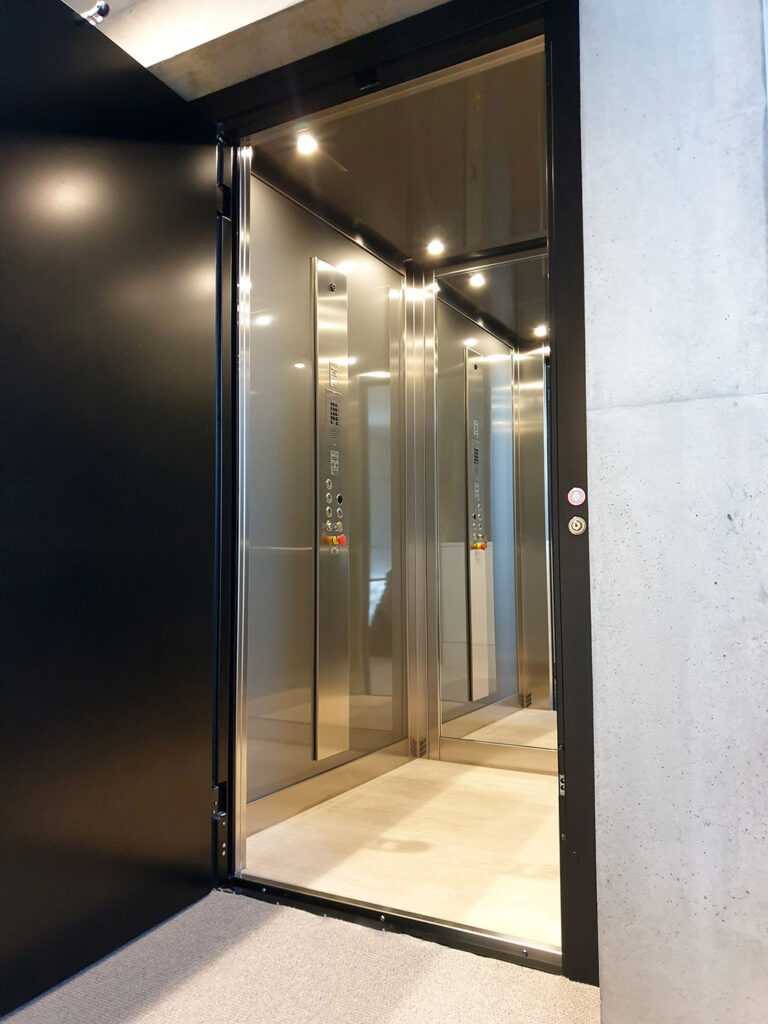 Open black landing door beside the mirrored stainless cabin of the E1 home lift with downlights, Toorak