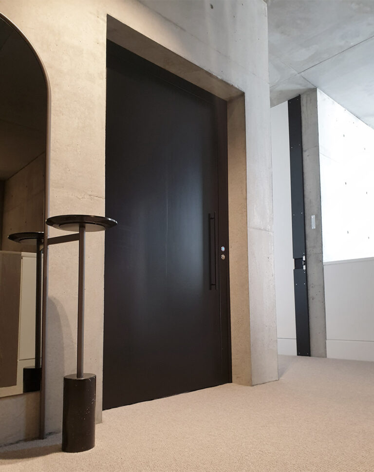 Closed dark timber lift door in a concrete wall beside a floor lamp