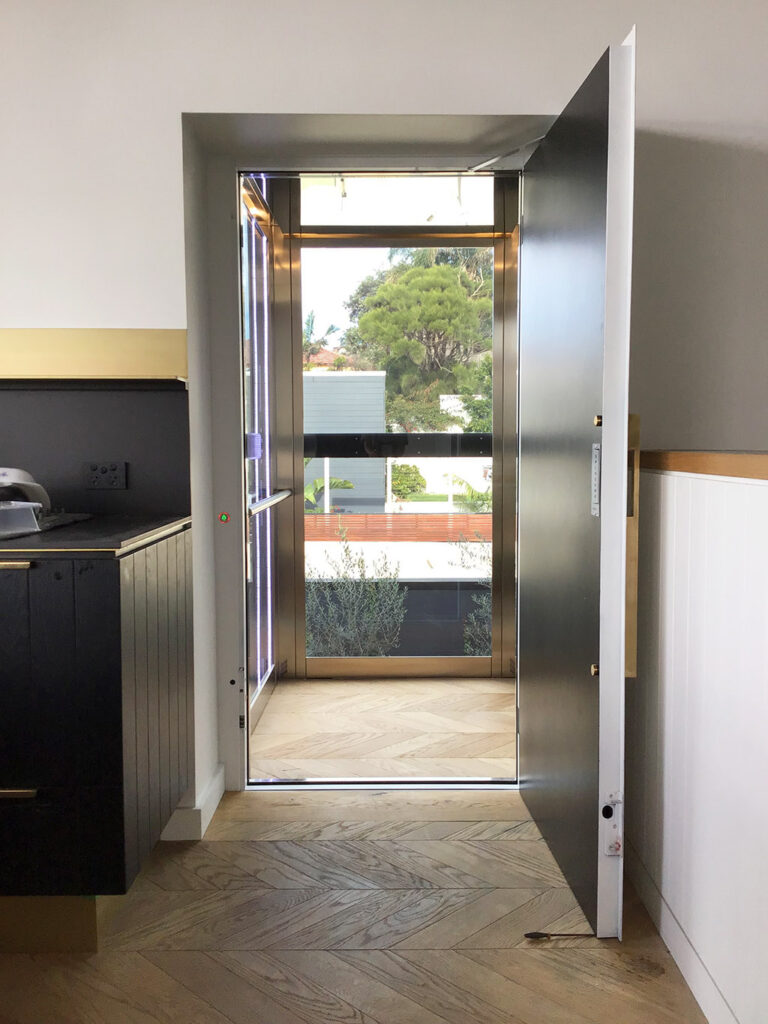 The swing door of the lift open from the kitchen, with the glass cabin and garden beyond