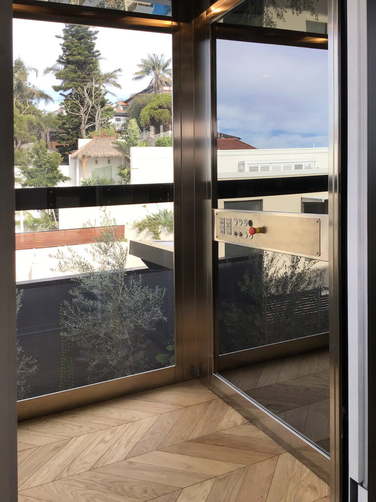 Inside the glass cabin with a stainless steel control panel, a chevron timber floor and views over Coogee rooftops