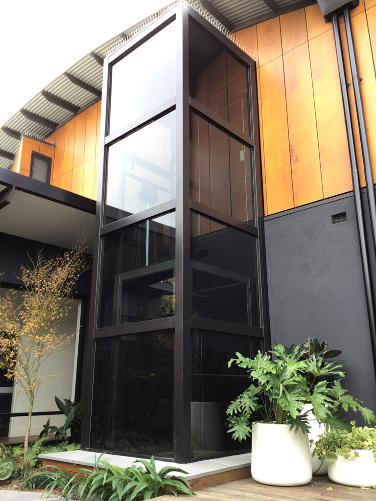 The black framed glass shaft against orange timber cladding and a charcoal wall, with planters beside a timber deck