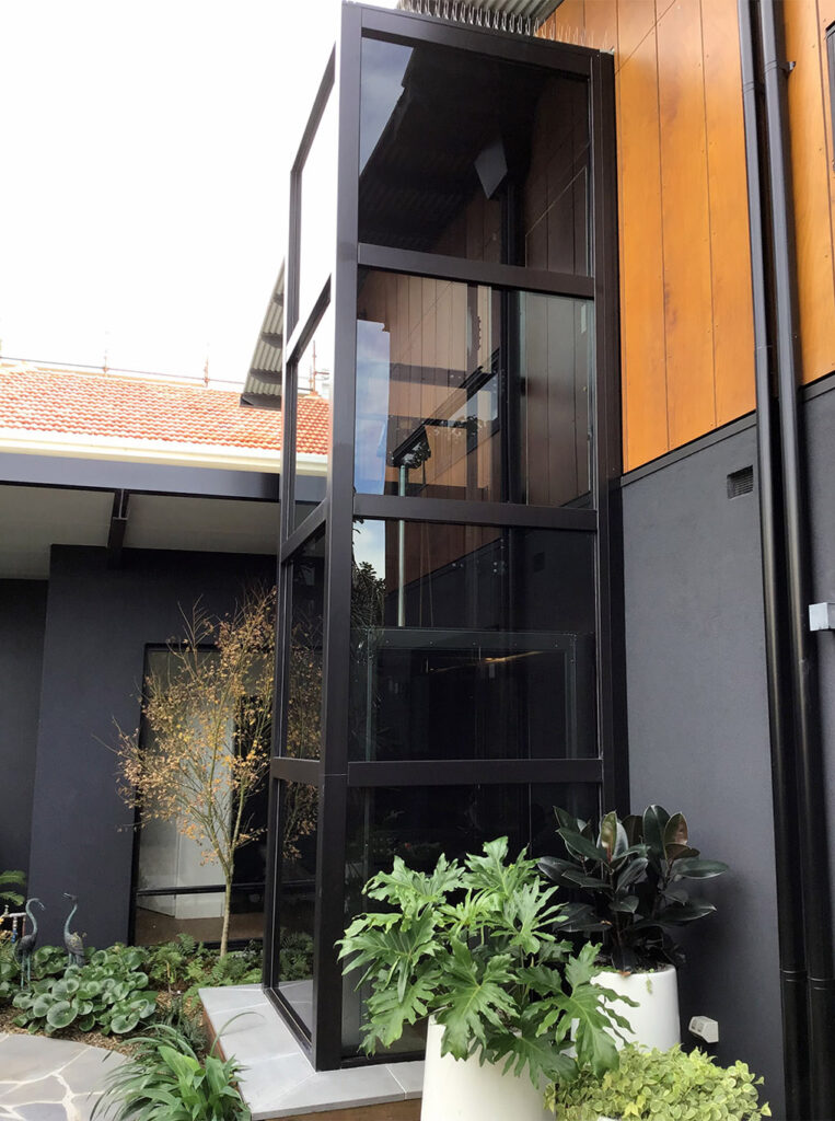 The black framed glass shaft rising beside timber cladding, with planting at its base