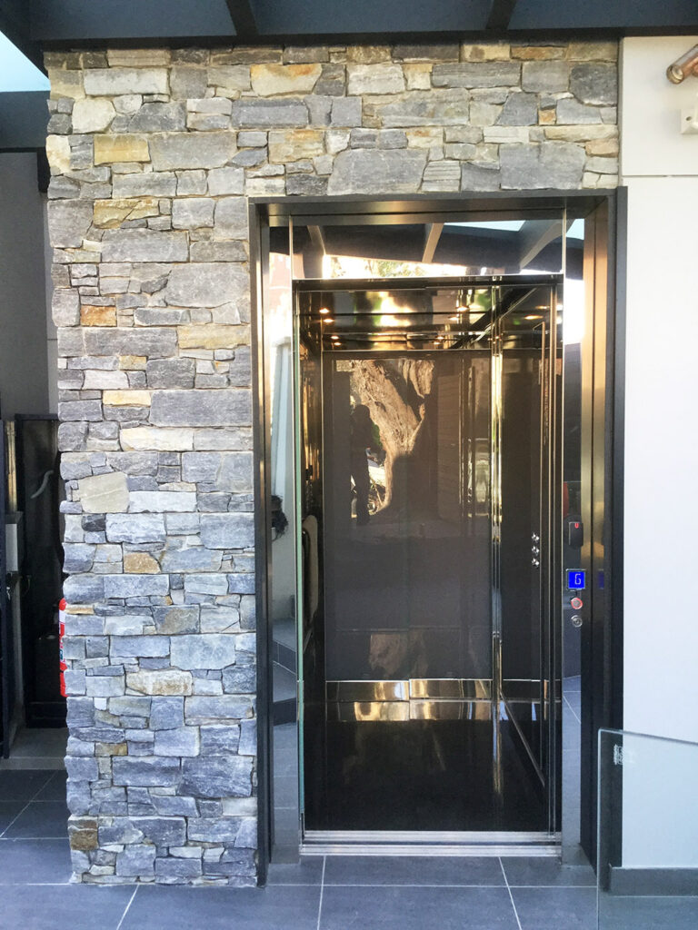 Home lift with its sliding door open in a blue stone shaft, showing the polished stainless cabin, in Fairlight