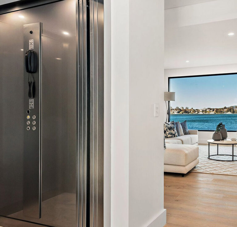 Stainless steel E2 home lift door and call panel beside a bright living room with water views