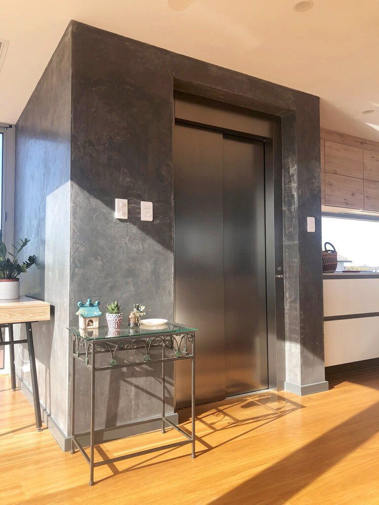Steel lift door in a dark textured surround beside a hall table and kitchen bench