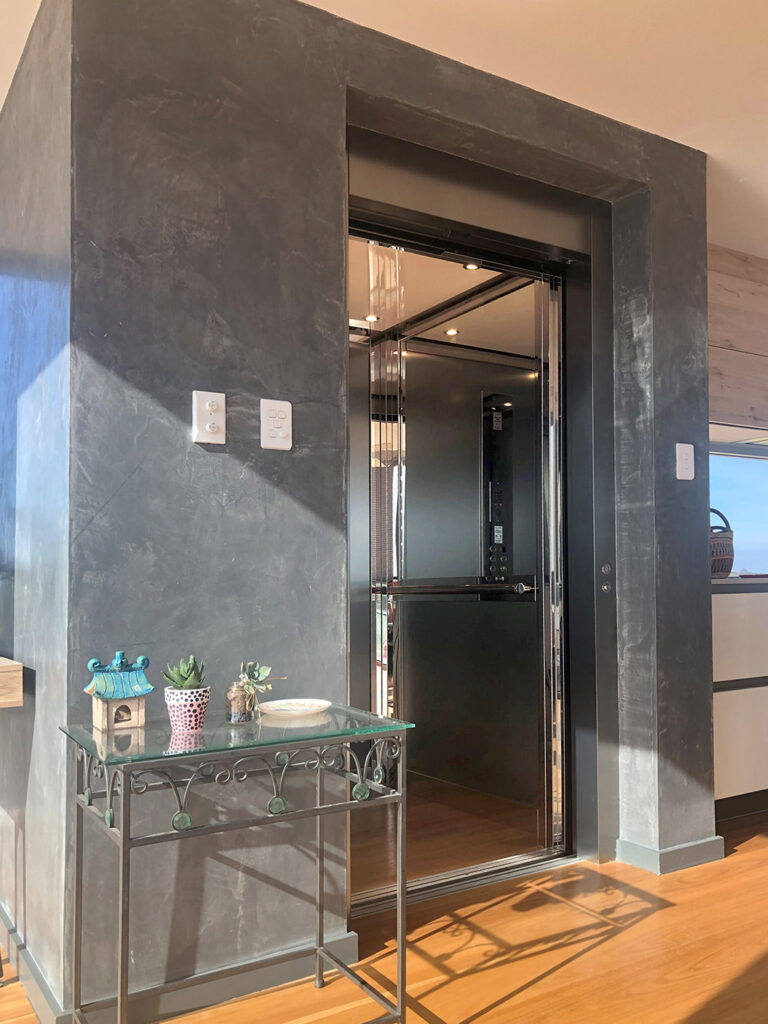 Open E2 lift with stainless steel doors in a dark textured wall surround beside a glass console table