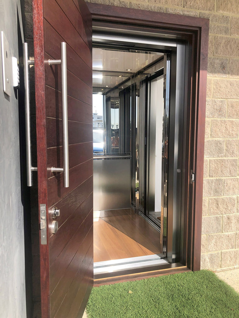 The lift seen through a timber front door with a long steel handle beside a brick wall