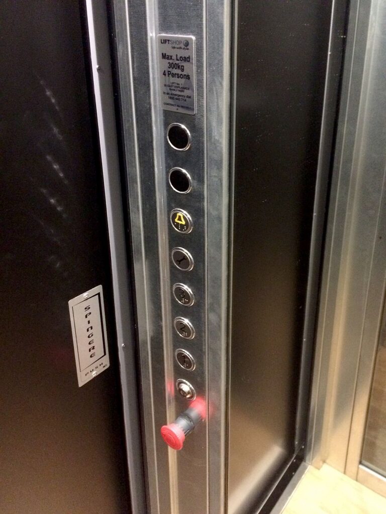 Lift control panel with round buttons for each floor and a red emergency button in the galvanized steel cabin
