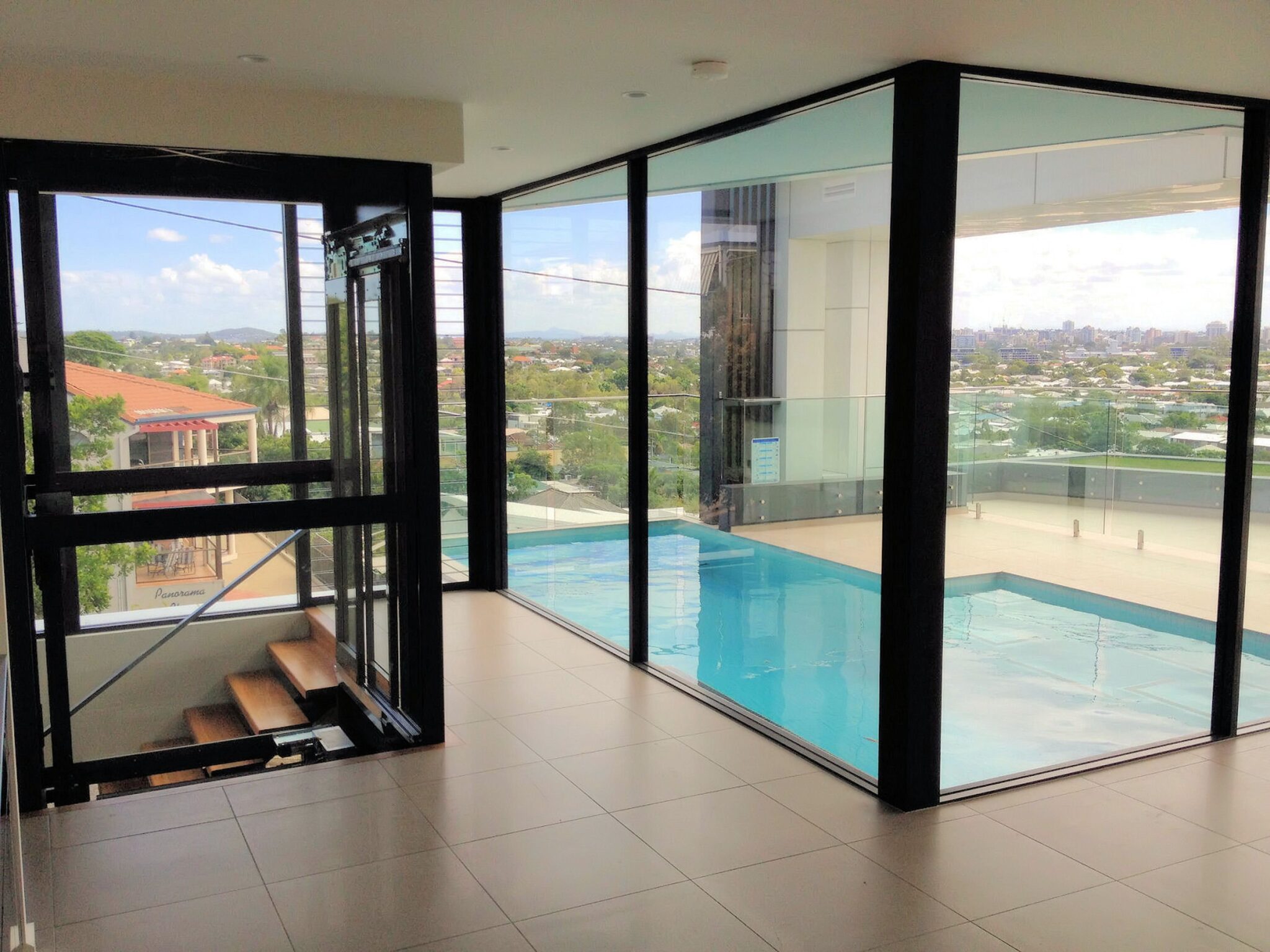 Glass-walled living area with a pool and city views beside the open glass lift shaft and timber stairs