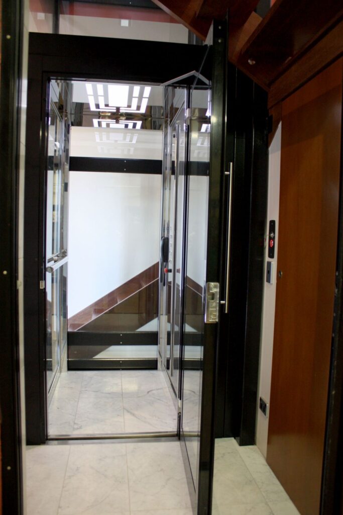 The black-framed glass lift door open onto a marble-floored landing, with the glass cabin and its ceiling light visible inside