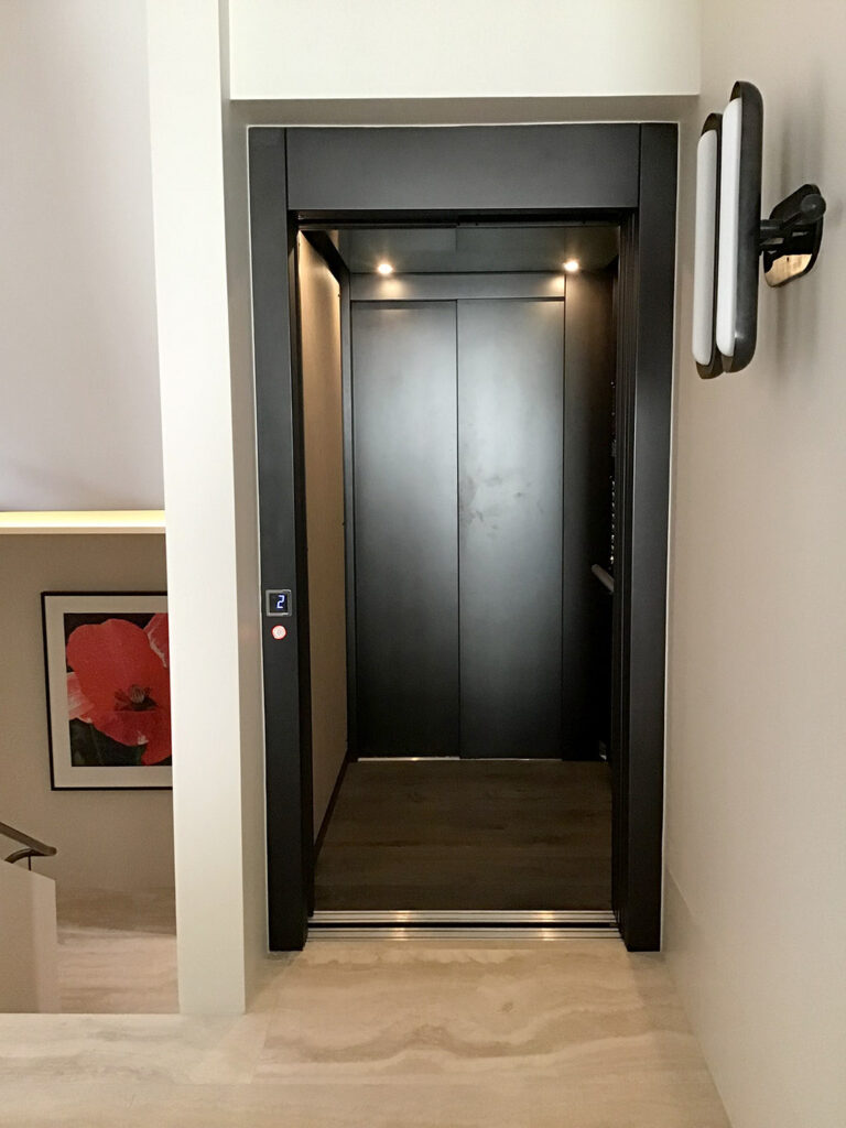 Open lift car with dark finishes and a floor indicator beside a stone-floored stairwell in a Bellevue Hill home