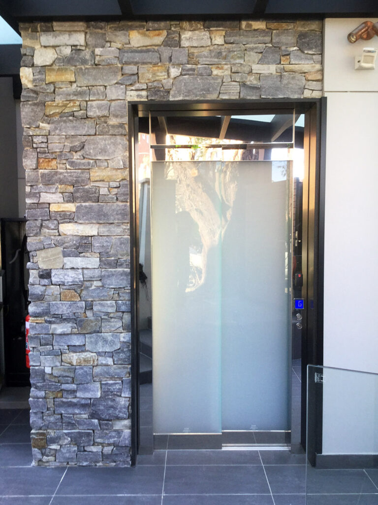 Frosted glass lift door framed by a stacked stone wall on a tiled floor