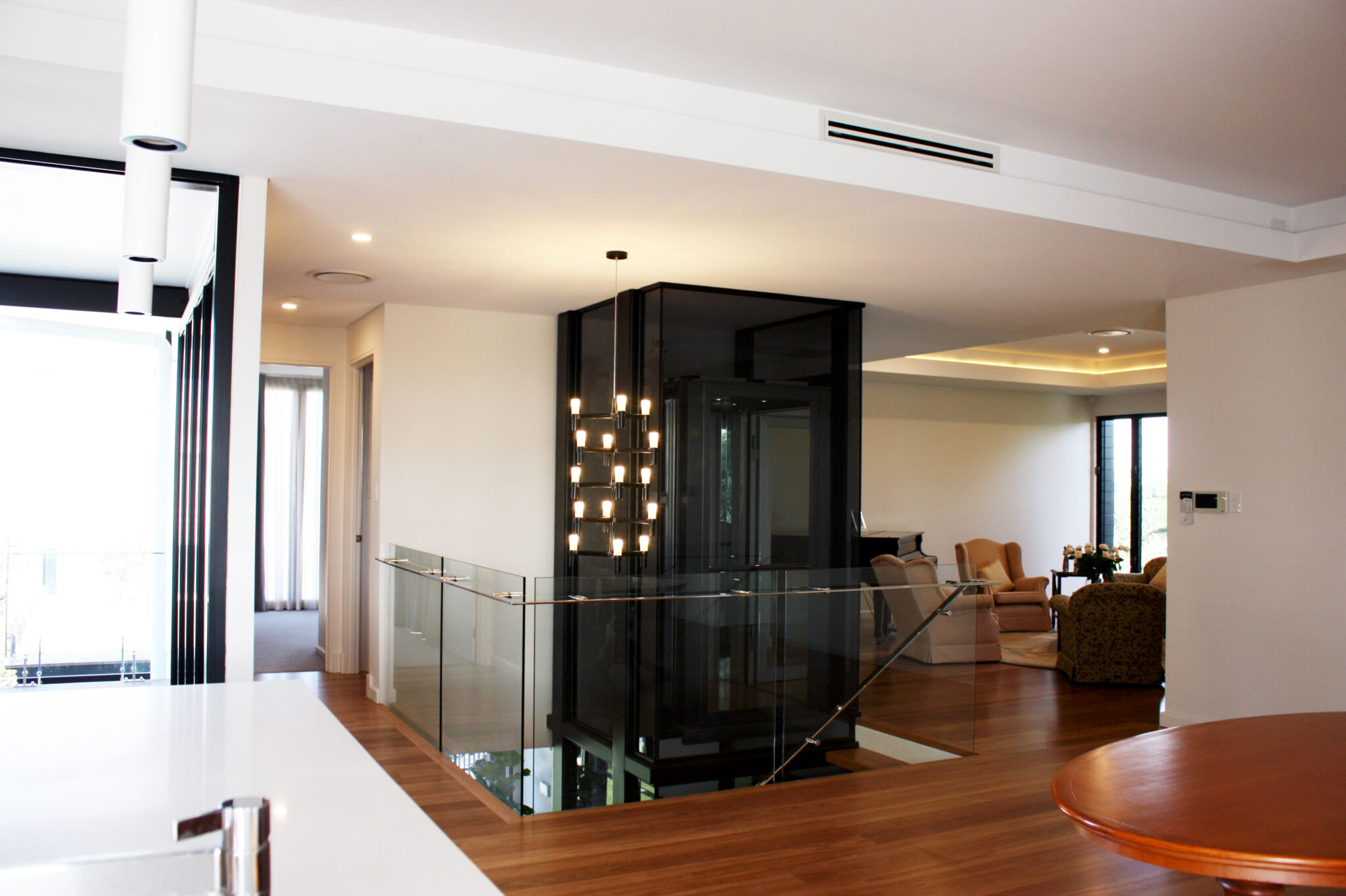 Black glass lift shaft with a chandelier beside a glass balustrade in an open-plan living area in Bulimba