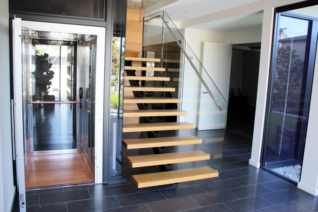 View through the open lift door across the garage-level landing to floating stairs and dark floor tiles