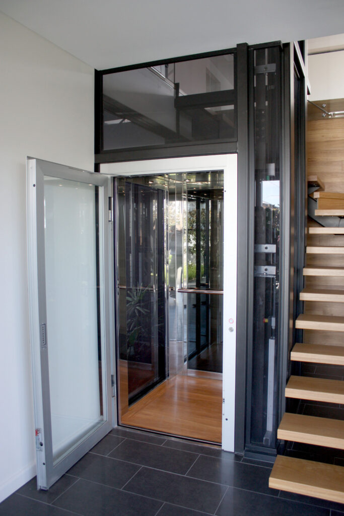 Open glass-panelled lift door on a black glass shaft beside a floating timber staircase