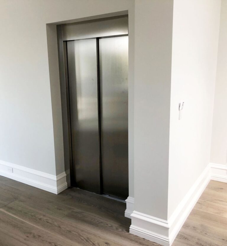 Closed brushed stainless steel lift doors beside white skirting and a timber floor