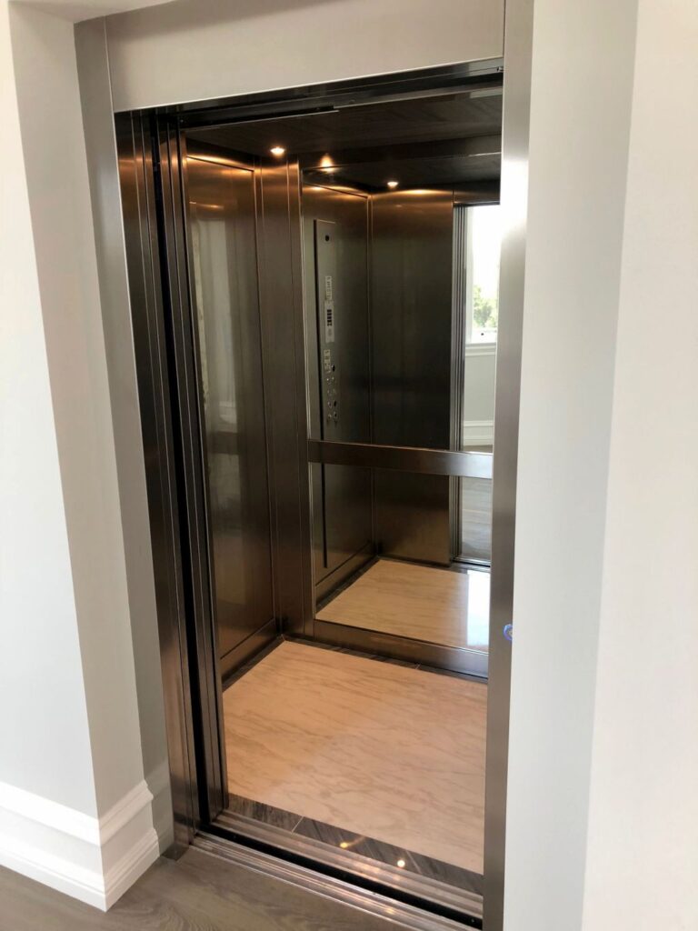 Brushed stainless steel lift door open to a cabin with a mirror rear wall and timber floor, seen from the hallway