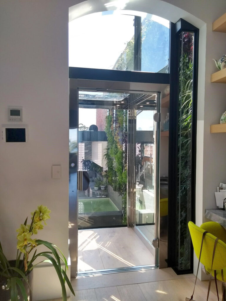 Glass fronted lift in a kitchen and dining area with an arched window above