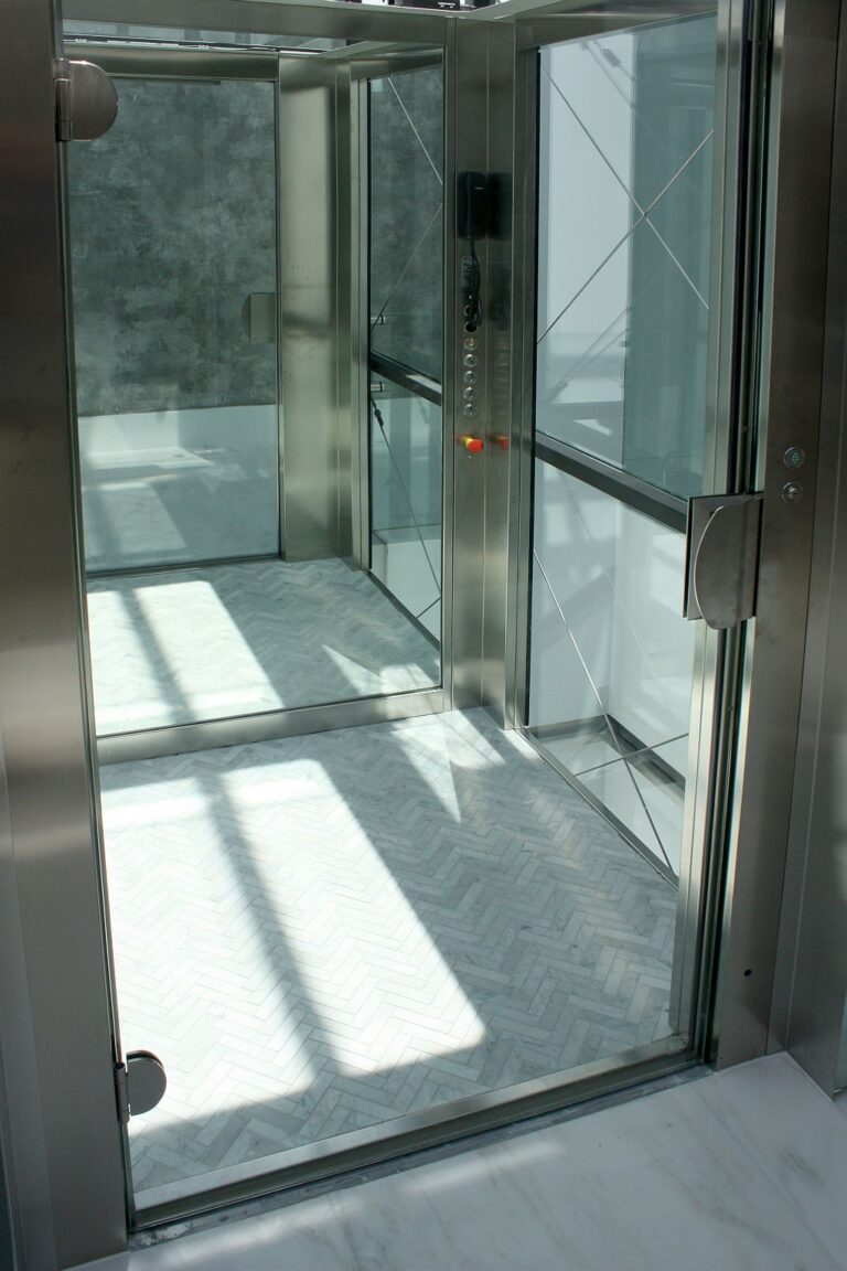 Inside the glass lift cabin with a tiled floor, a stainless steel control panel and sunlight through the glass